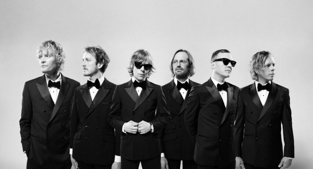 A group of men in black suits and sunglasses posing against a light background.